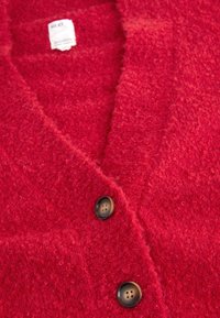 Cardigan rouge texturé avec deux boutons ronds foncés et une étiquette de marque visible près du col.