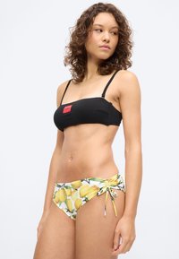Γυναίκα που φοράει μαύρο μπαντό μπικίνι τοπ με κόκκινο λογότυπο και λευκά μπικίνι σλιπ με κίτρινο σχέδιο λεμονιού και δεσίματα στο πλαϊνό μέρος.