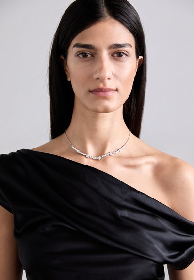 Collana d'argento con accenti di cristallo scintillanti, caratterizzata da un design delicato che mette in risalto la clavicola, indossata sopra un top in tessuto nero drappeggiato.