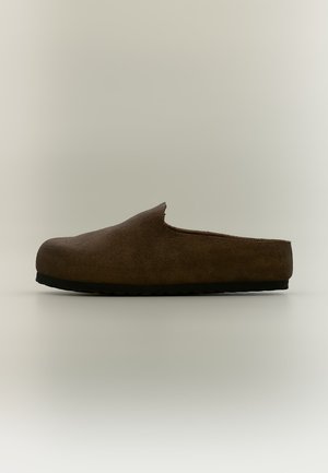 Scarpa slip-on in pelle scamosciata marrone con suola nera, tacco basso e punta arrotondata, mostrata di profilo su sfondo neutro.