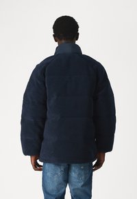 Marineblå fleecejakke med polstret design, brede ermer, høy krage og teksturert overflate, kombinert med lyseblå jeans.