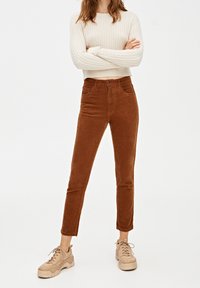 Personne portant un pull court crème en maille torsadée, un pantalon en velours côtelé marron taille haute, et des baskets beiges épaisses, debout les bras croisés.