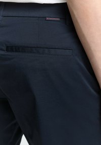 Jūras spēku bikses ar gludu auduma tekstūru, kas aprīkotas ar aizmugurējo kabatu un mazu sarkanu zīmola etiķeti, kas izšūta uz jostas.