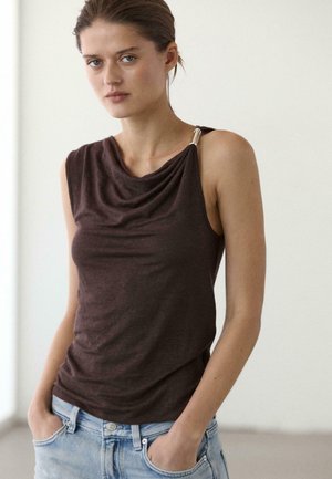 Junge Frau mit zurückgebundenen Haaren, die ein dunkles, ärmelloses Oberteil mit gerafftem Ausschnitt und hellblaue Jeans trägt, mit den Händen in den Taschen stehend.