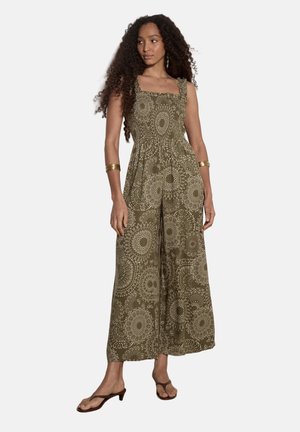 Femme portant une combinaison pantalon large kaki avec des motifs circulaires complexes, accessoirisée de bracelets et boucles d’oreilles dorés, posant pieds nus.