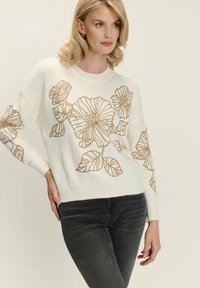 Eine blonde Frau trägt einen weißen Pullover mit großen beigen Blumenumrissen und dunkelgrauen Jeans und posiert mit einer Hand auf der Hüfte.