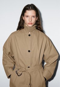 Bruine oversized jas met hoge kraag, zwarte knopen, afneembare riem en een losse pasvorm. De stof is glad en textuurrijk.