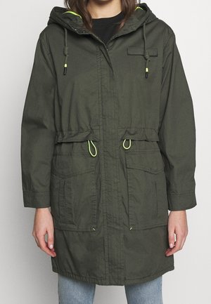 Femme portant un parka à capuche vert olive avec poches avant et cordons de serrage jaune fluo, associée à un jean bleu clair.