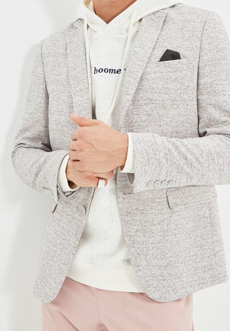 Blazer grigio chiaro strutturato con due bottoni, indossato sopra un hoodie bianco. Presenta un fazzoletto da taschino nero e pantaloni rosa chiaro.