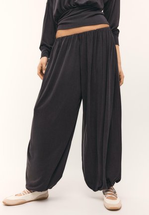 Personne portant un pantalon sarouel ample gris foncé avec une ceinture élastique et des chaussures blanches à lacets, debout devant un fond clair.