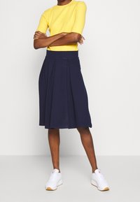 Keltainen ribattukangas toppi yhdistettynä tummansiniseen laskostettuun hameeseen, jossa on istuva vyötärö ja aaltoileva siluetti. Valkoiset tennarit viimeistelevät asun.