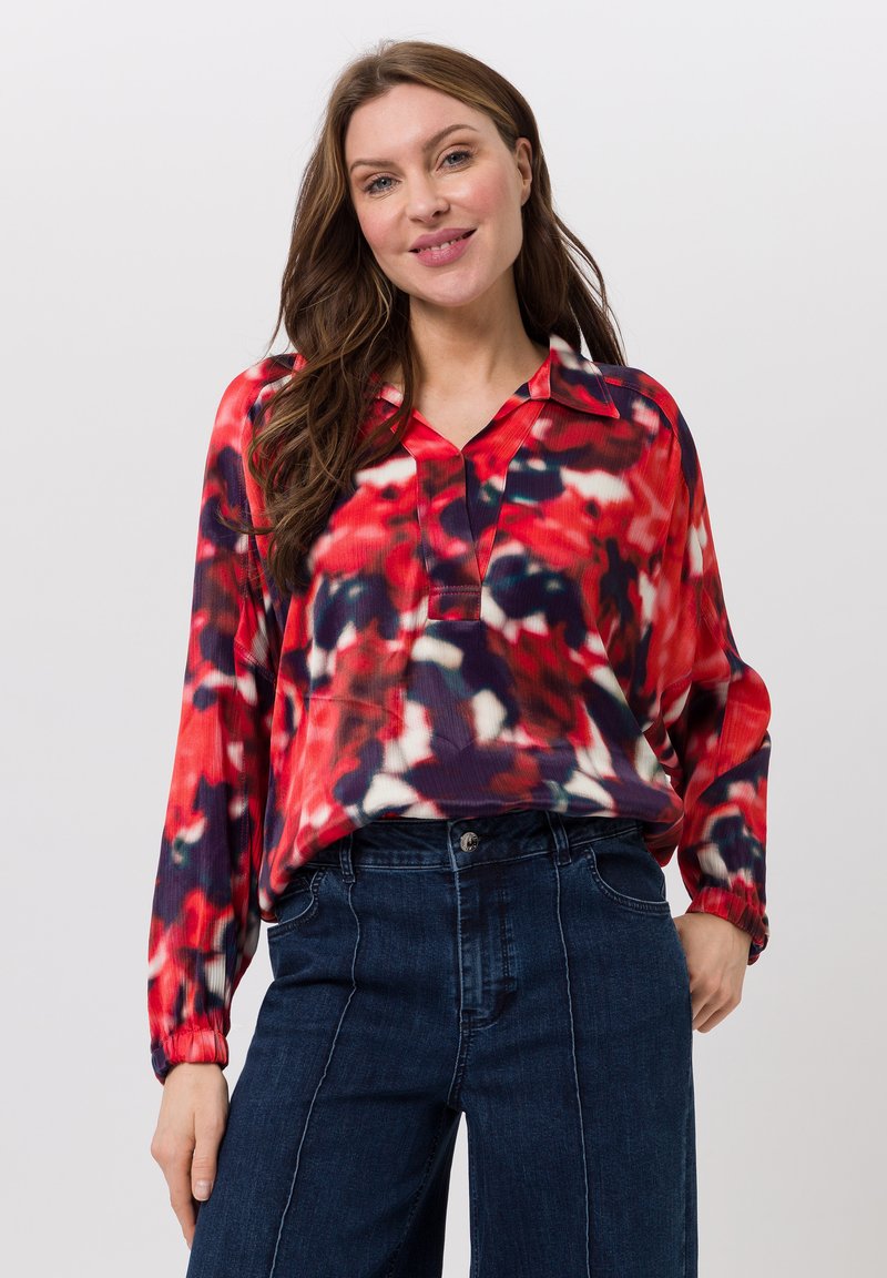 Vrouw met lang bruin haar in een rood, zwart en wit geblokt blouse en donkerblauwe hoge taille jeans, glimlachend met een hand in de zak.