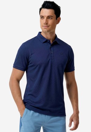 Uomo che indossa una polo blu navy e pantaloni azzurri, in piedi con le mani in tasca, guardando leggermente di lato.