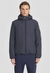 Marineblauwe waterdichte jas met capuchon, ritsafsluiting, elastische manchetten, twee zijzakken en een klein logo op de mouw.