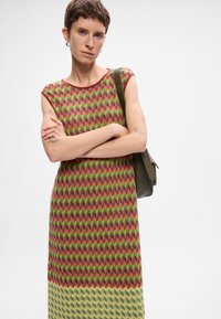 Άτομο με κοντά μαλλιά που φοράει ένα αμάνικο φόρεμα σε μορφή ζιγκ-ζαγκ σε πράσινο, κόκκινο και κίτρινο χρώμα, κρατώντας μια σκούρα πράσινη τσάντα ώμου.