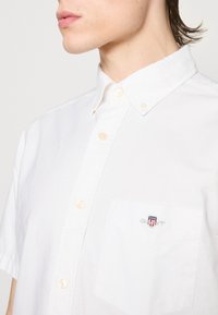 Zbliżenie osoby noszącej białą koszulę z krótkim rękawem, zapinaną na guziki, z ostrym kołnierzykiem i małym logo na kieszeni piersiowej.