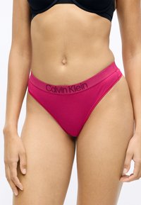 Różowa bawełniana stringi Calvin Klein z kontrastującym czarnym paskiem z logo, o gładkiej teksturze i klasycznym kroju majtek.