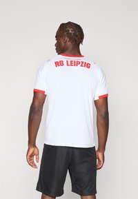 Bijela sportska majica s crvenim akcentima, na leđima s natpisom "RB LEIPZIG" u podebljanom crvenom slovima, uparena s crnim šortsem.