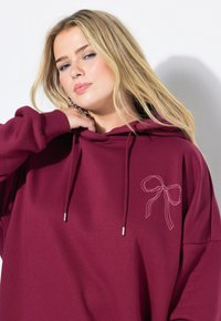Felpa bordeaux con tasca frontale, coulisse regolabile e grafica di un fiocco bianco nella zona sinistra del petto. Tessuto morbido e forma rilassata.