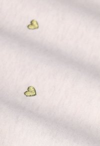 Tissu rose clair avec deux petits cœurs en or brodés. La texture semble douce et lisse, mettant en avant des détails de design minimalistes.