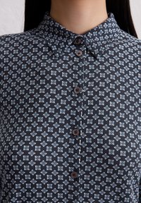 Bluse mit Kragen aus marineblauem Stoff mit einem geometrischen Muster in Weiß und Blau. Sechs braune Knöpfe schließen die vordere Blende.