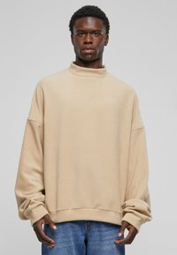 Beige fleecegenser med høy krage, løs passform, senkede skuldre og ribbestrikkede mansjetter, kombinert med blå denim jeans.