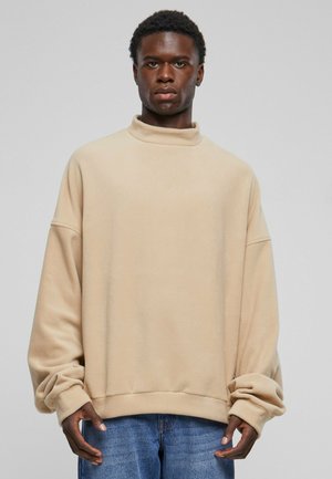 Beige fleece sweatshirt with a high collar, loose fit, dropped shoulders, and ribbed cuffs, paired with blue denim jeans.