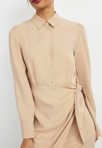 Abito beige a maniche lunghe con colletto a camicia, patta frontale e dettaglio in vita annodato. Tessuto liscio con una leggera lucentezza.