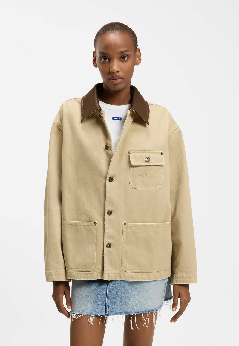 Donna che indossa una giacca beige con colletto marrone sopra una camicia bianca e una gonna di denim sfrangiata, in piedi davanti a uno sfondo bianco neutro.