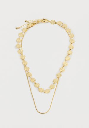 Collar de oro de doble capa con una cadena que presenta pequeños encantos en forma de flores y una cadena delgada y lisa sobre fondo blanco.