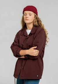 Bordowa lekka kurtka z klasycznym kołnierzem, zapięciem na guziki i elastycznymi mankietami. Nosi się ją na białej koszuli, w połączeniu z niebieskimi dżinsami.