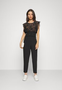 Black lace top with cap sleeves, gathered waist, and high-waisted black trousers. Model holds a small beige clutch and wears white loafers.