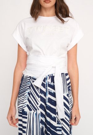Kobieta ubrana w białą koszulkę Twinset Milano z podwiniętymi rękawami i paskiem do wiązania, w połączeniu z niebiesko-białymi spodniami w abstrakcyjne paski.