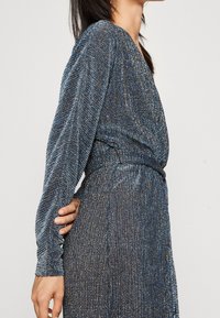 Robe à manches longues en bleu métallique et argenté, présentant un design portefeuille, un tissu texturé avec des plis verticaux et une taille cintrée.