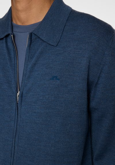 Uomo che indossa un cardigan in maglia blu con zip sopra una maglietta blu a girocollo, che mostra il colletto e la parte superiore del petto.