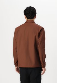 Giacca leggera marrone con colletto, polsini elasticizzati e una texture liscia. Design semplice, senza motivi visibili o decorazioni.