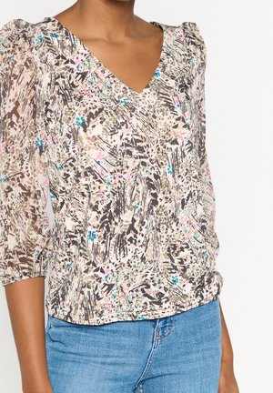 Femme portant un chemisier à col en V avec un motif abstrait multicolore et des manches trois-quarts bouffantes, associé à un jean en denim bleu.