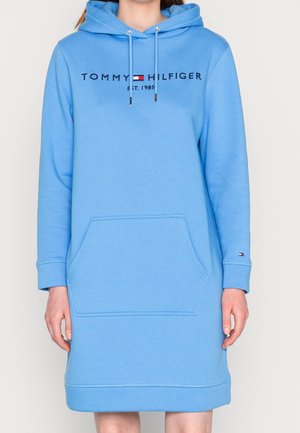 Femme portant une robe sweat à capuche Tommy Hilfiger bleu clair avec une poche avant et un texte logo sur la poitrine.
