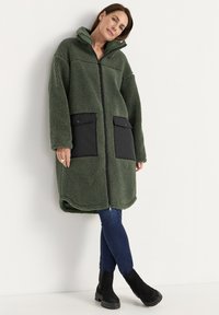 Vrouw in een lange groene sherpajas met grote zwarte zakken, blauwe jeans en zwarte enkellaarsjes, staand tegen een witte muur.