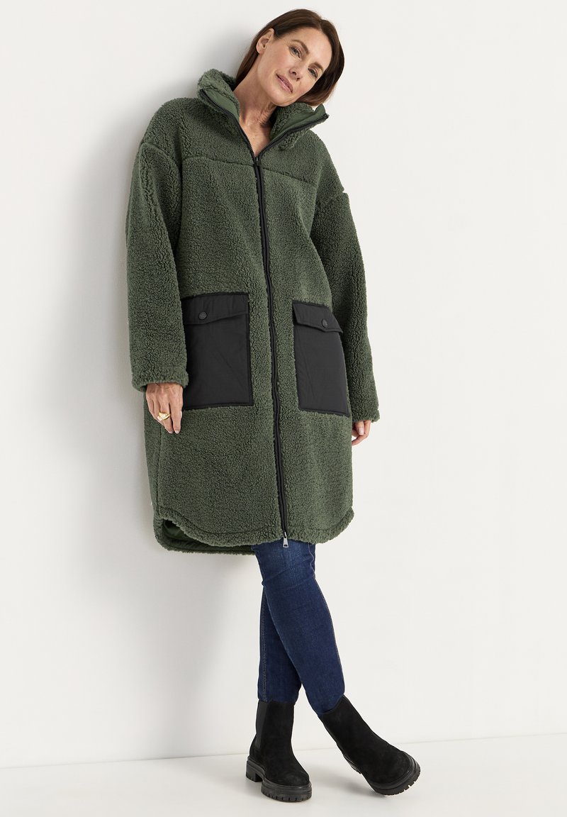 Vrouw in een lange groene sherpajas met grote zwarte zakken, blauwe jeans en zwarte enkellaarsjes, staand tegen een witte muur.