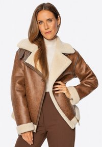 Braune Kunstlederjacke mit cremefarbenem Lammfellbesatz, breitem Kragen, Reißverschluss und dekorativen Riemen auf den Schultern. Strukturierte Oberfläche.