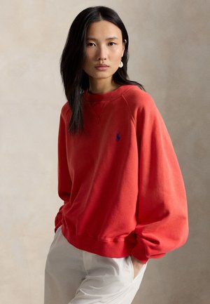 Noor naine, kellel on pikad tumedad juuksed, punane dressipluus ja valged püksid, seisab ühe käega taskus neutraalse tausta ees.