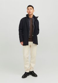 Chaqueta exterior azul marino con un cuello alto y un forro interior acolchado gris, presenta botones a presión y una forma no estructurada. Combinada con pantalones claros.
