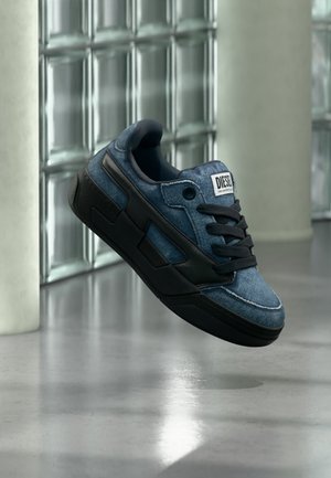 Kék és fekete Diesel sportcipő fekete fűzőkkel, amely lebeg egy szürke padló felett, üvegtömb fal és fehér oszlopok közelében.