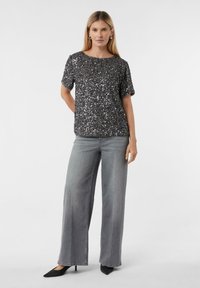 Schwarzes, kurzärmeliges Oberteil mit Pailletten und rundem Ausschnitt, kombiniert mit weit geschnittenen grauen Jeans und schwarzen, spitz zulaufenden High Heels. Glatte Stoffstruktur.