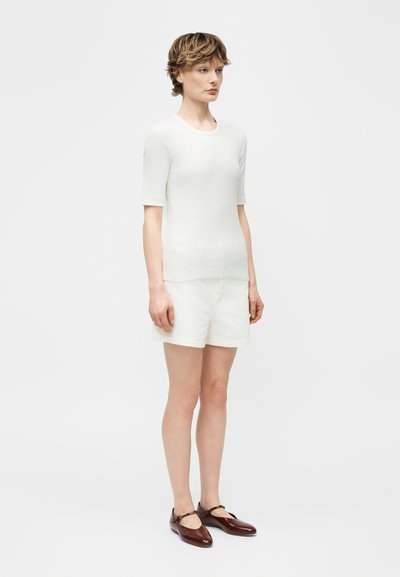 Νεαρό άτομο με κοντά μαλλιά που φοράει λευκό κοντομάνικο μπλουζάκι, λευκά σορτς και σκούρα καφέ παπούτσια Mary Jane, στέκεται μπροστά σε απλό φόντο.