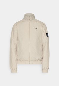 Beżowa kurtka zapinana na zamek z wysokim kołnierzem, elastycznymi mankietami i dołem, kieszeniami z przodu, logo "cK" na piersi oraz naszywką na lewym rękawie.