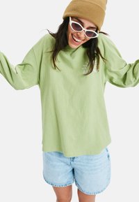 Jasnozielona koszula z długim rękawem w luźnym kroju, z małym logo. Połączona z niebieskimi szortami dżinsowymi, tworzy swobodny wygląd.