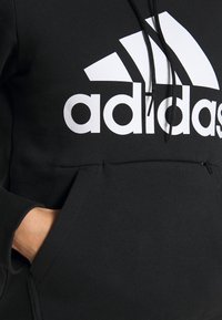 Černá mikina s kapucí se velkým bílým logem Adidas na přední straně, obsahující klokankovou kapsu a černé šňůrky na kapuci. Měkká textura.
