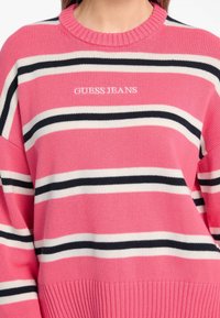 Rosa Strickpullover mit horizontalen Streifen in Weiß und Schwarz, Rundhalsausschnitt und geripptem Saum mit "GUESS JEANS" Logo auf der Vorderseite.